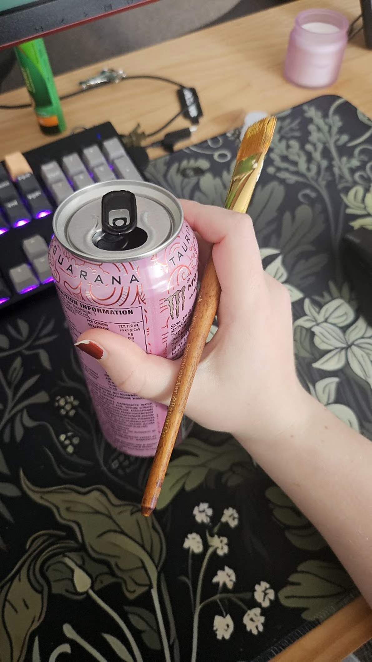 A paint brush and a pink Monster can in one hand, over the floral desk mat